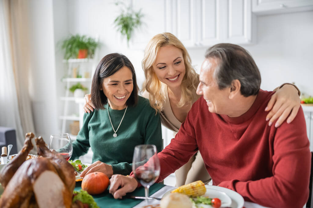 Позитивная женщина обнимает многонациональных родителей возле ужина в честь Дня Благодарения дома  - Фото, изображение