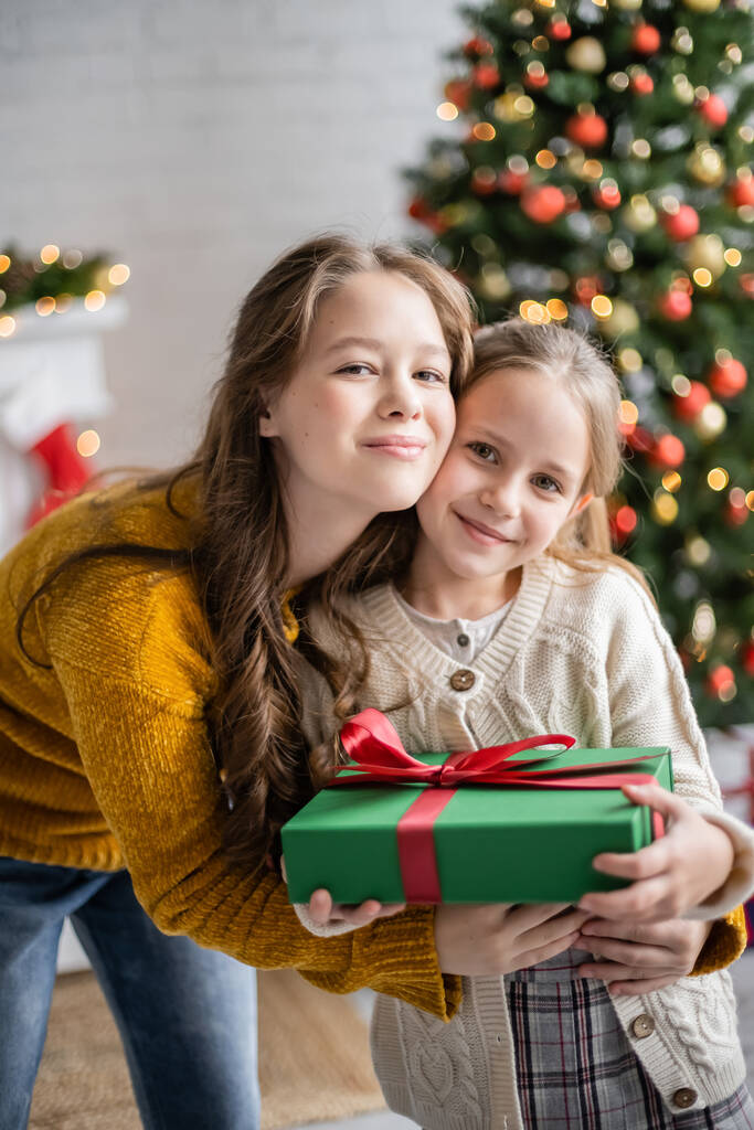glimlachende tiener knuffelen zus met heden tijdens kerst thuis  - Foto, afbeelding