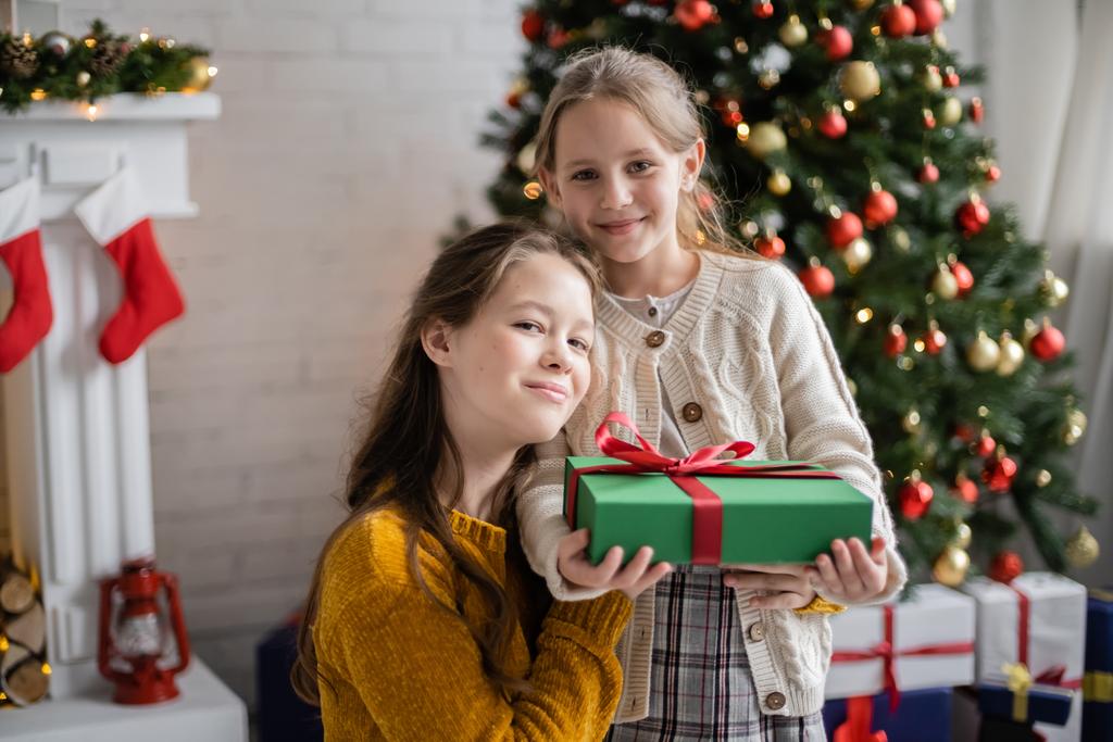 gelukkig zussen met geschenk doos kijken naar camera in de buurt versierde kerstboom op wazig achtergrond - Foto, afbeelding