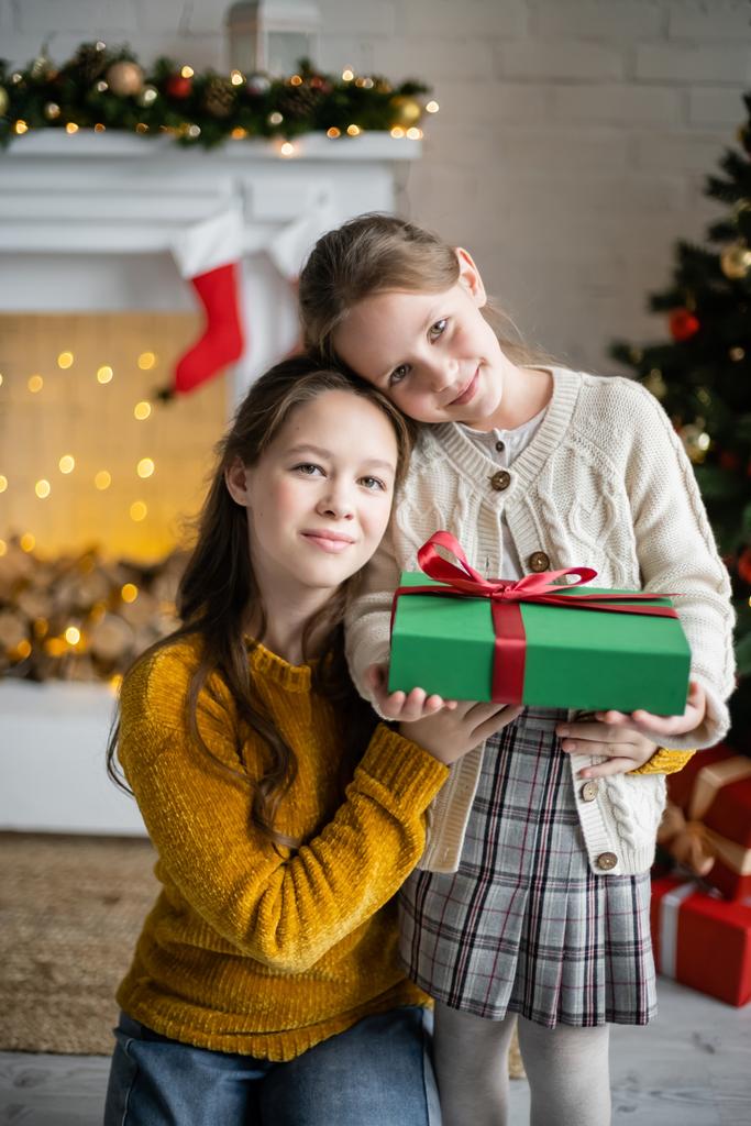 улыбающиеся сестры держат подарочную коробку и смотрят на камеру в гостиной с рождественским декором - Фото, изображение