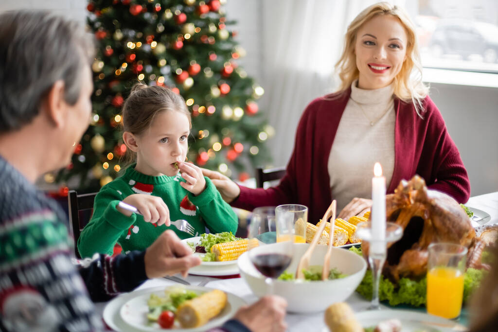 dívka s vidličkou s vánoční večeře v blízkosti rozmazané dědeček a usmívající se maminka - Fotografie, Obrázek