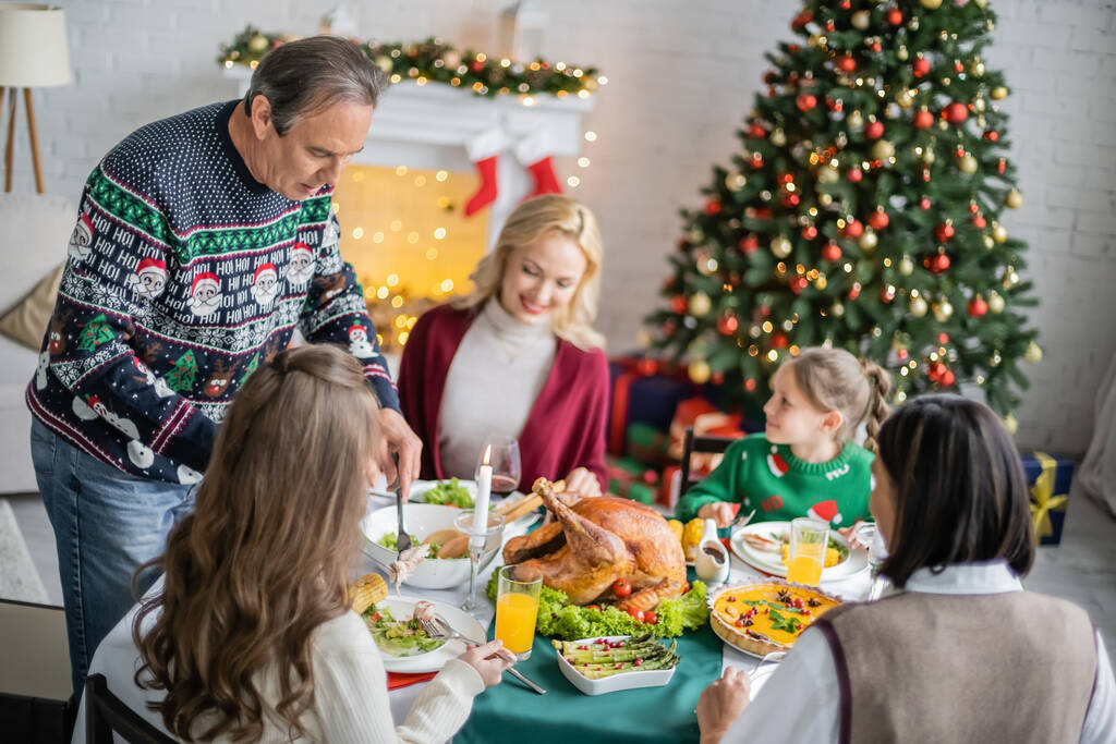 senior man serving vegetable salad near granddaughter and multiracial family during christmas dinner - Photo, Image