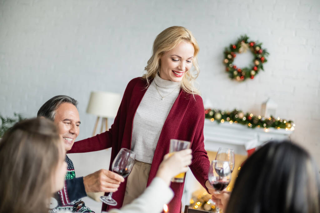mujer alegre de pie cerca de tostadas familiares con copas de vino mientras celebra la Navidad en casa - Foto, Imagen