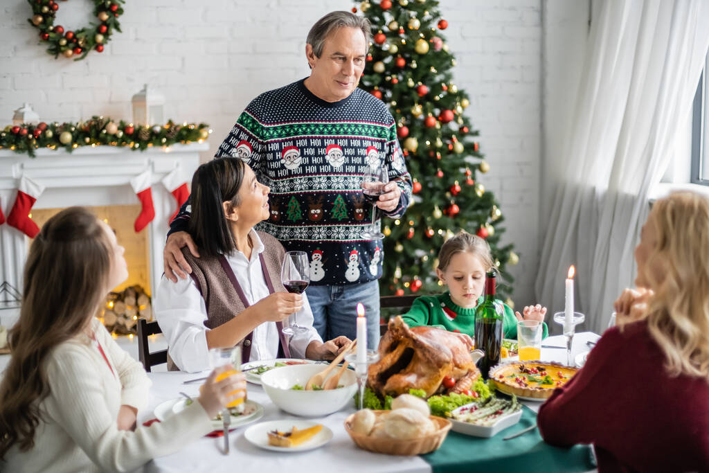 homem sênior brindar com copo de vinho tinto perto de família multiétnica durante o jantar de Natal festivo - Foto, Imagem