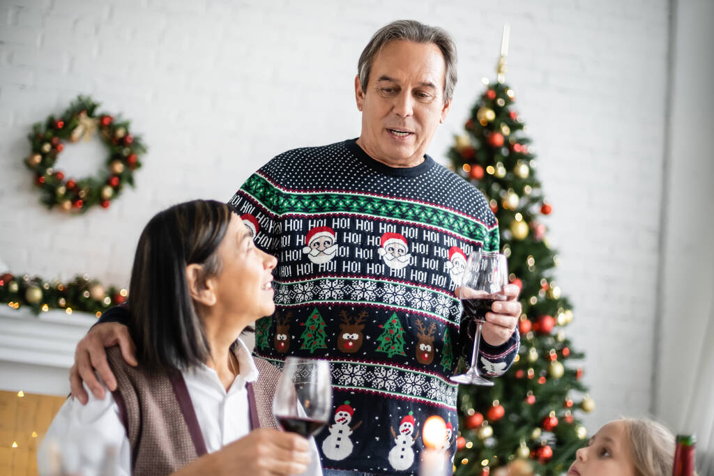 senior man hugging multiracial wife while toasting with glass of red wine near granddaughter during christmas dinner - Photo, Image
