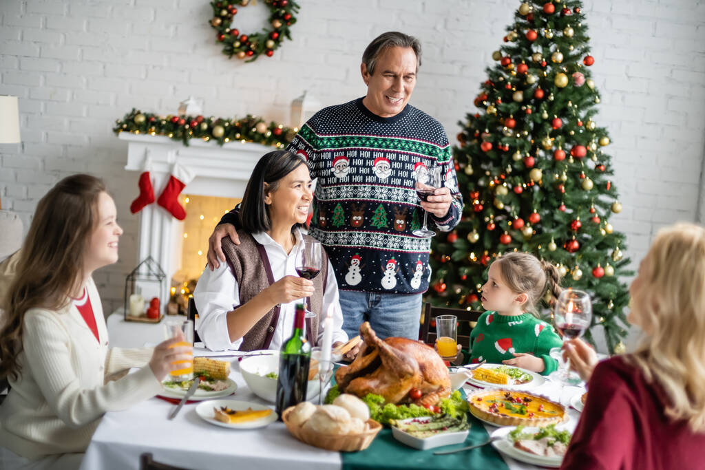 hombre sonriente tostadas con copa de vino tinto durante la cena festiva con la familia interracial cerca del árbol de Navidad decorado - Foto, imagen
