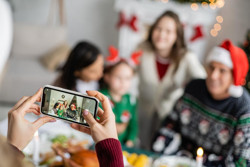 mulher com smartphone tirando fotos de família interracial desfocada celebrando o Natal em casa - Foto, Imagem