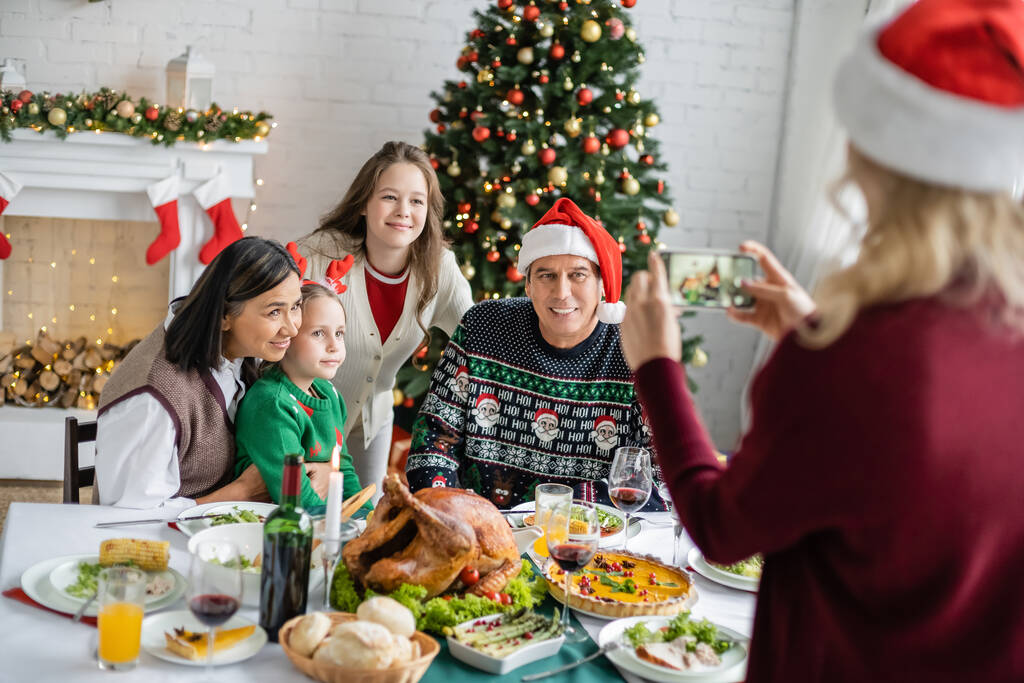 verschwommene Frau fotografiert glückliche interrassische Familie in der Nähe von festlichem Abendessen und Weihnachtsbaum - Foto, Bild