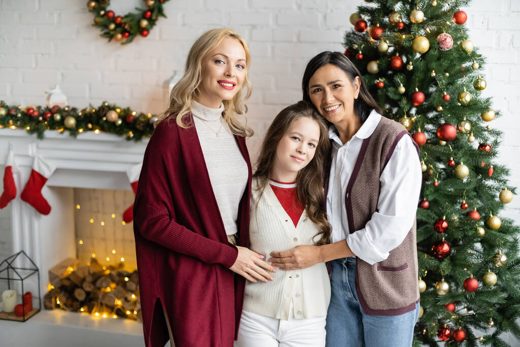 felice donna bionda guardando la macchina fotografica vicino figlia e mamma multirazziale in soggiorno con decorazioni natalizie - Foto, immagini