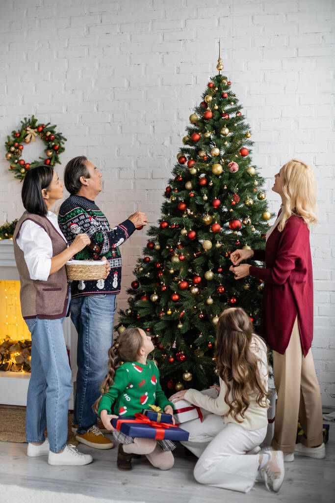 ギフトボックスの近くの床に座って異人種間の家族を見てクリスマスツリーを飾る - 写真・画像