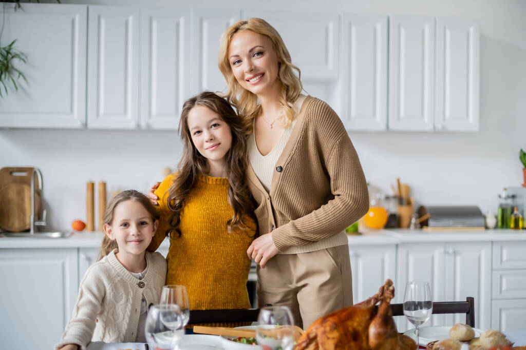 blondi nainen tyttärien kanssa hymyilee kameralle lähellä kiitospäivää illallinen tarjoillaan keittiössä - Valokuva, kuva