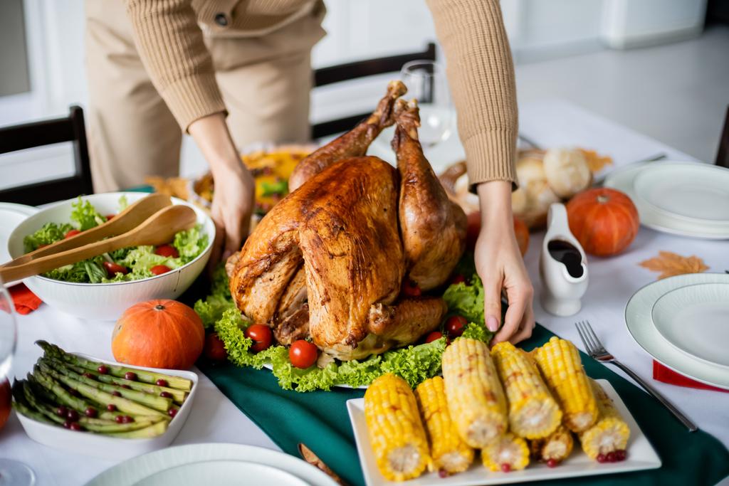 祝いの感謝祭のディナーのためにグリルコーンと野菜サラダの近くに伝統的な七面鳥を提供する女性の部分的なビュー - 写真・画像