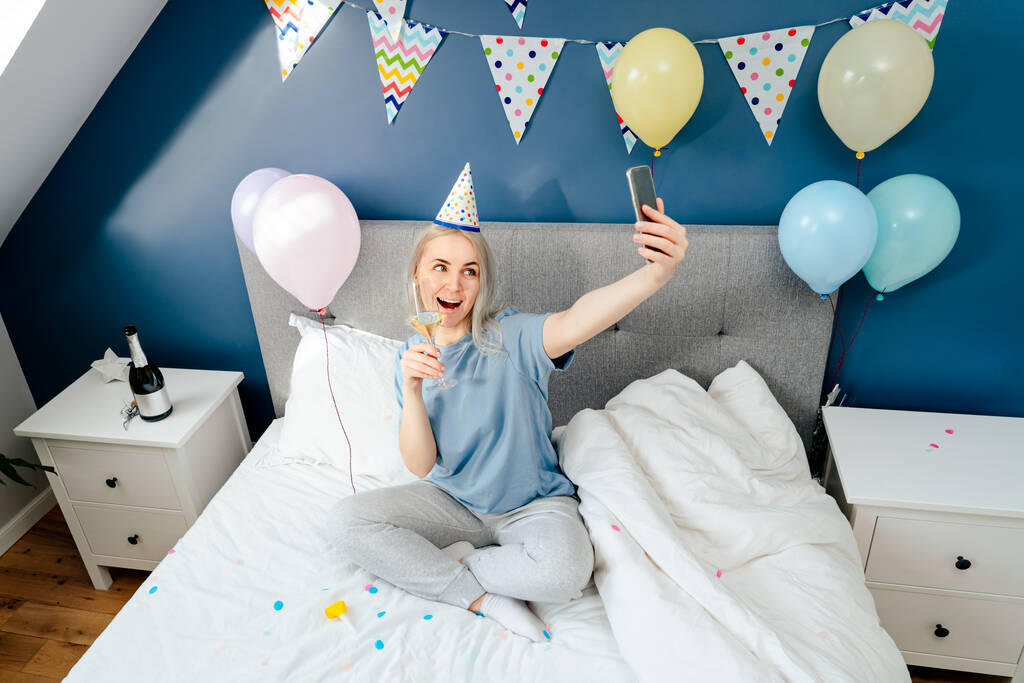 寝室でシャンパンと風船でベッドの上に座っている間、パジャマとパーティーキャップで幸せな感情的な女性は、誕生日の朝に携帯電話で自撮りを行います。誕生日おめでとう. - 写真・画像