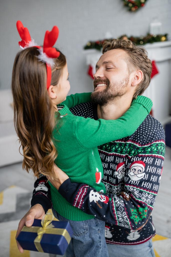 Uśmiechnięty mężczyzna w swetrze przytula córkę i trzyma świąteczny prezent w domu  - Zdjęcie, obraz