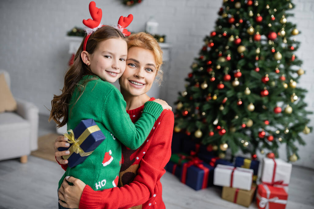 glimlachende vrouw in trui knuffelen dochter en het houden van kerstcadeau thuis  - Foto, afbeelding
