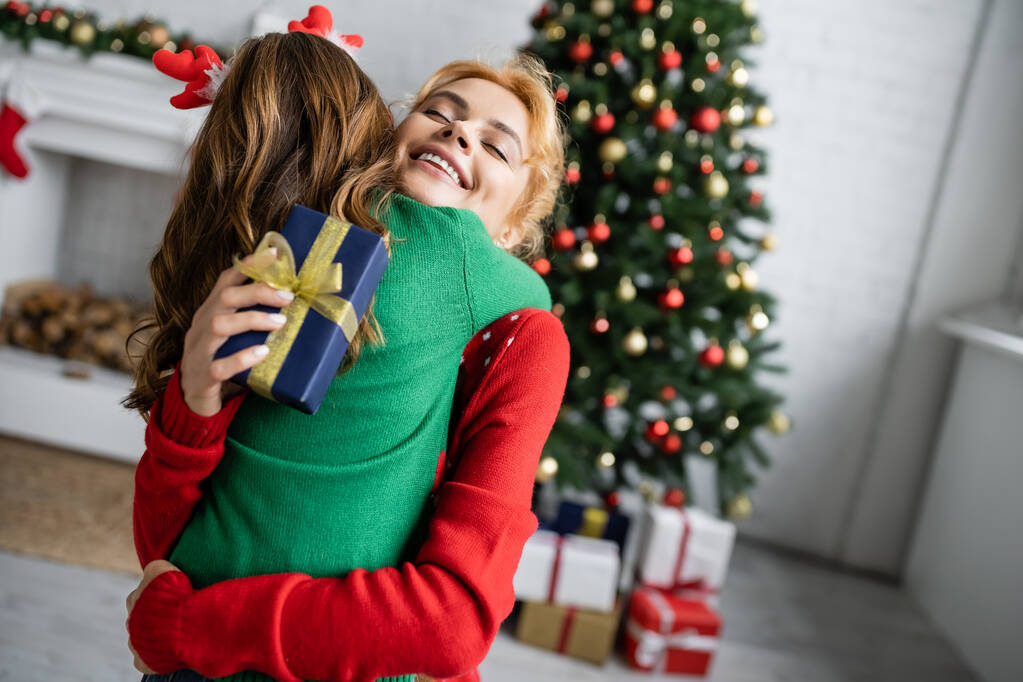 Улыбающаяся мама в свитере обнимает дочь и держит новогодний подарок дома  - Фото, изображение