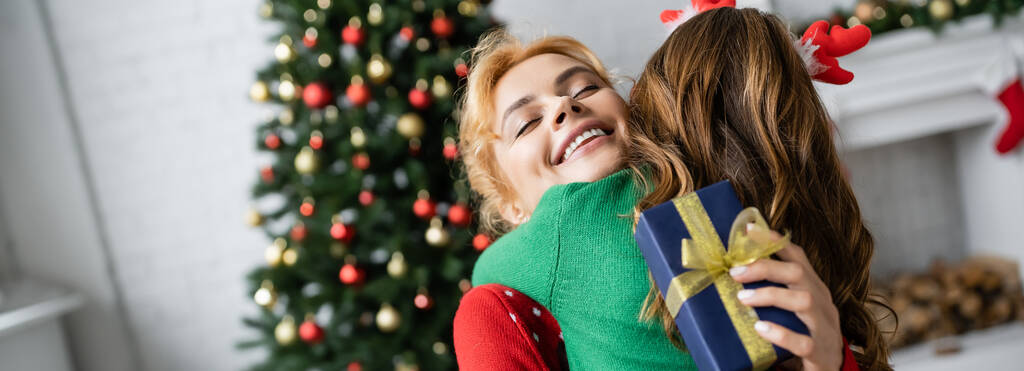 Usmívající se rodič objímající dceru a držící vánoční dárkovou krabici doma, prapor  - Fotografie, Obrázek