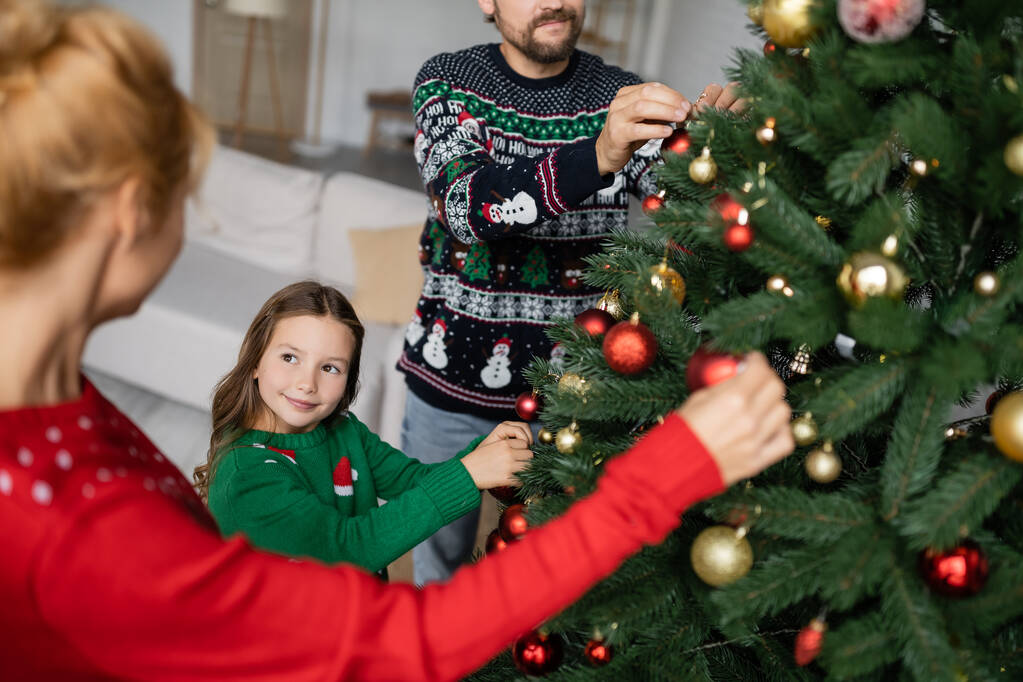 Enfant souriant en pull décorant l'arbre de Noël avec les parents à la maison  - Photo, image