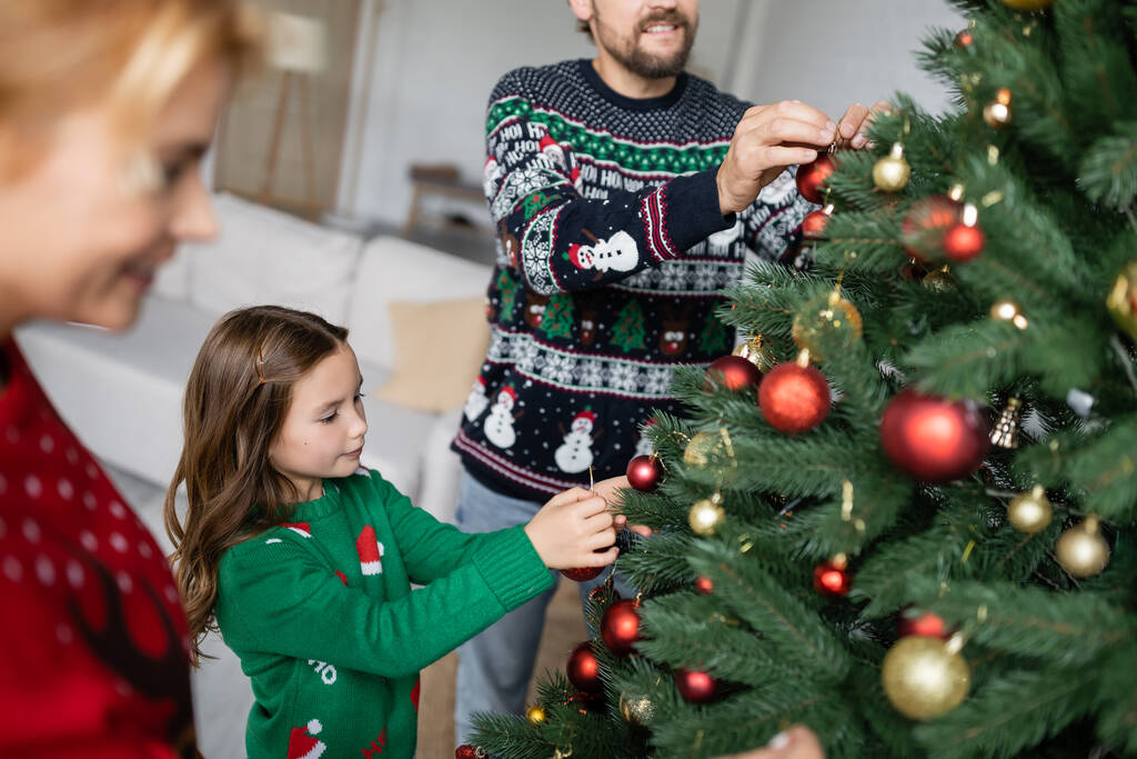 Dívka zdobení vánoční stromek s kuličkami v blízkosti rozmazané rodiče doma  - Fotografie, Obrázek