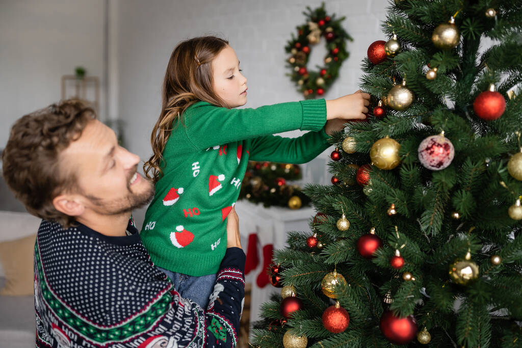Sonriente hombre sosteniendo hija decorando árbol de Navidad en casa  - Foto, imagen