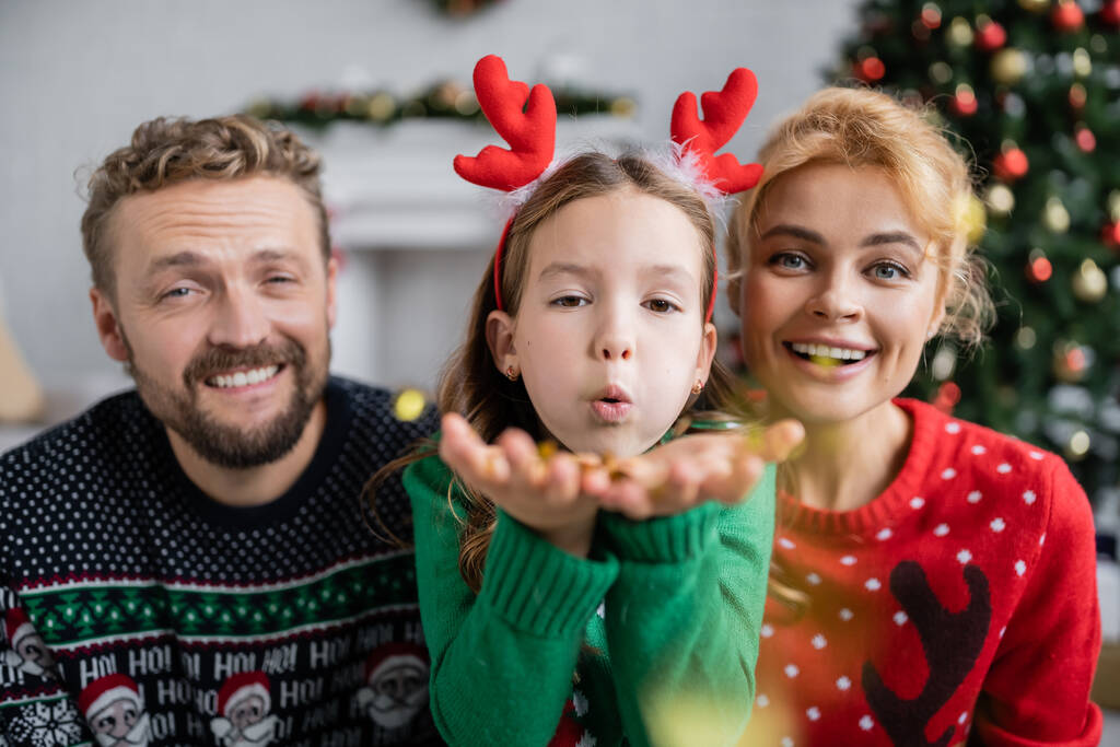 Lány fúj konfetti a kamera közelében mosolygós szülők karácsonyi pulóverek otthon  - Fotó, kép