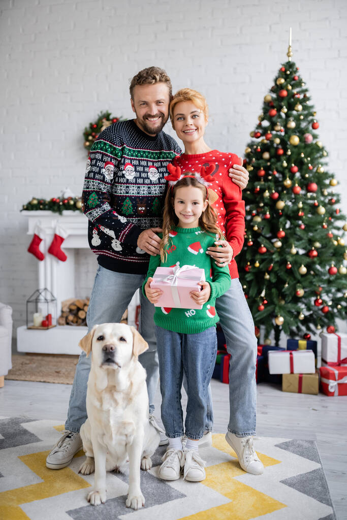 Позитивная семья обнимает дочь с подарком на Рождество и лабрадором дома  - Фото, изображение