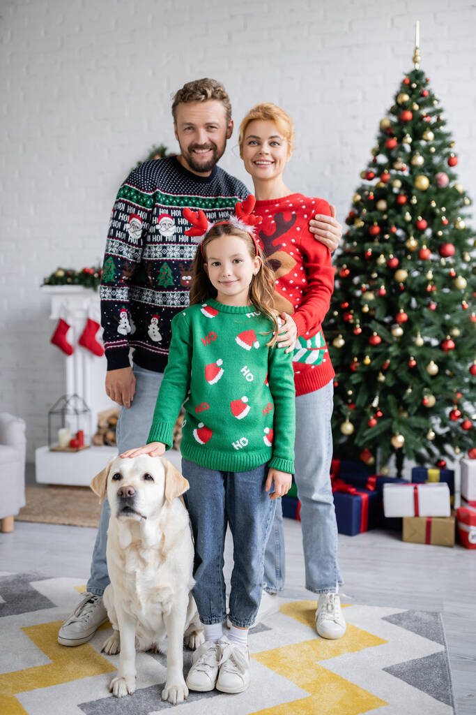 Gülümseyen ebeveynler, Labrador 'un yanında kızlarına sarılıyorlar ve evde bulanık Noel ağacı görüyorlar.  - Fotoğraf, Görsel