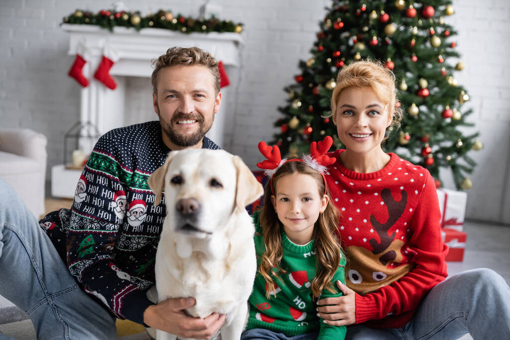 Wesoła rodzina w ciepłych swetrach przytula rozmazanego labradora i patrzy w kamerę w domu  - Zdjęcie, obraz