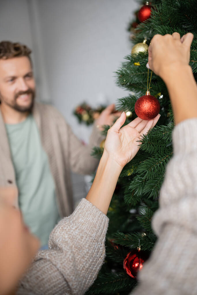 木の近くにクリスマスボールを持っている女性とぼやけた夫で家に  - 写真・画像
