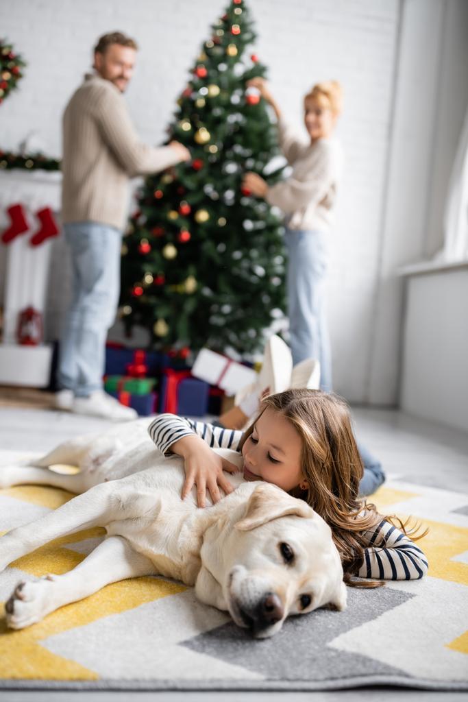 Улыбающаяся девушка обнимает лабрадора возле зарытых родителей и рождественской елки дома  - Фото, изображение