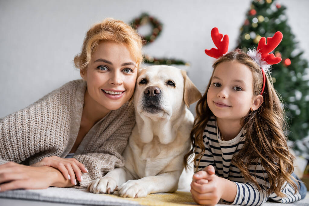 Pozytywne mama i córka z opaską świąteczną patrząc na aparat w pobliżu labrador na podłodze w domu  - Zdjęcie, obraz