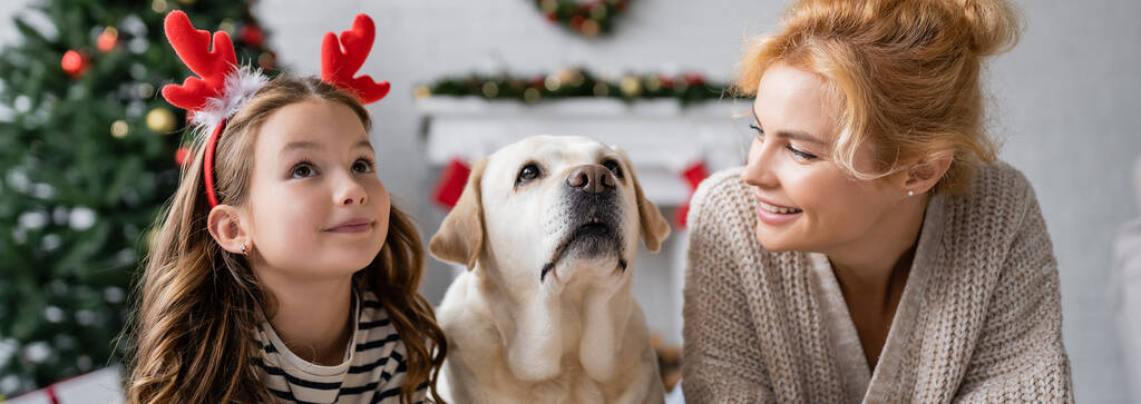 Positive Frau betrachtet Kind mit Weihnachts-Stirnband bei Labrador zu Hause, Banner  - Foto, Bild