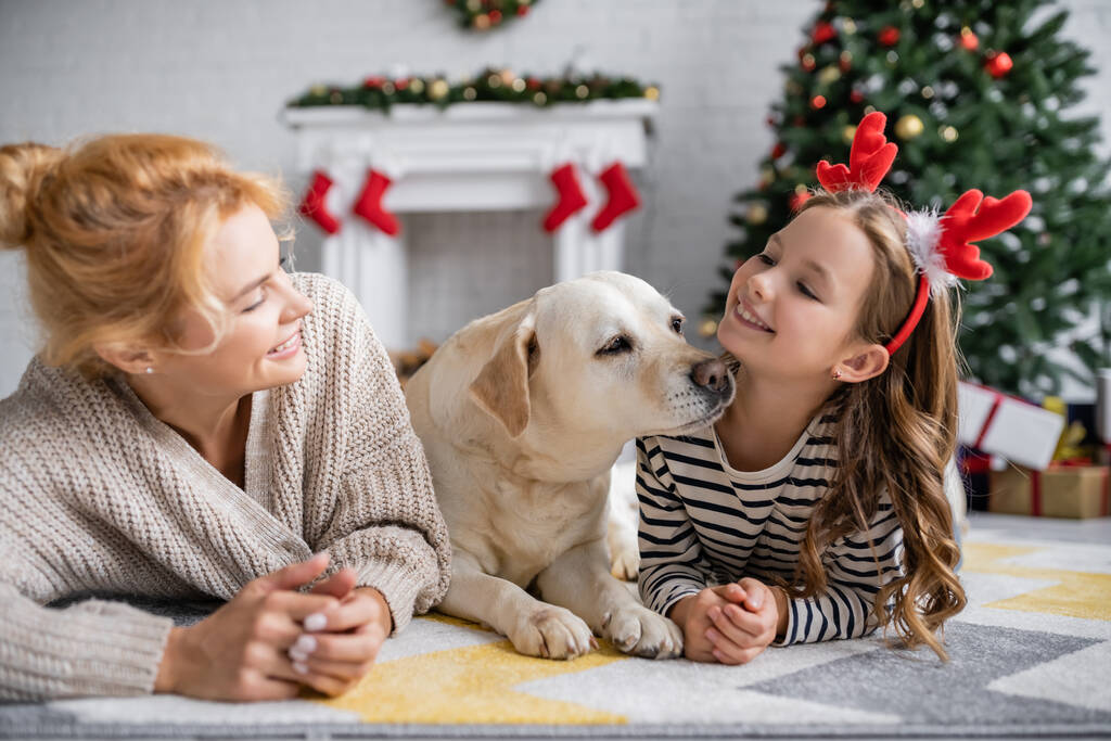 Sonriente madre mirando a su hija con diadema navideña y labrador en casa  - Foto, imagen