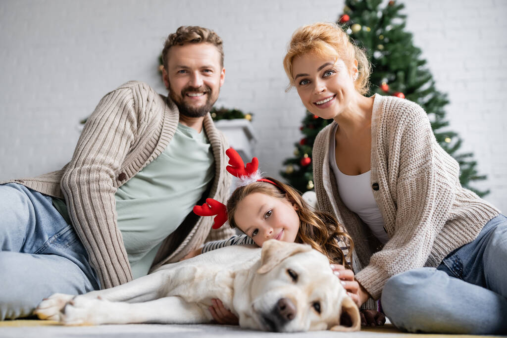 Positieve ouders kijken naar camera in de buurt dochter met kerst hoofdband en labrador op de vloer thuis  - Foto, afbeelding