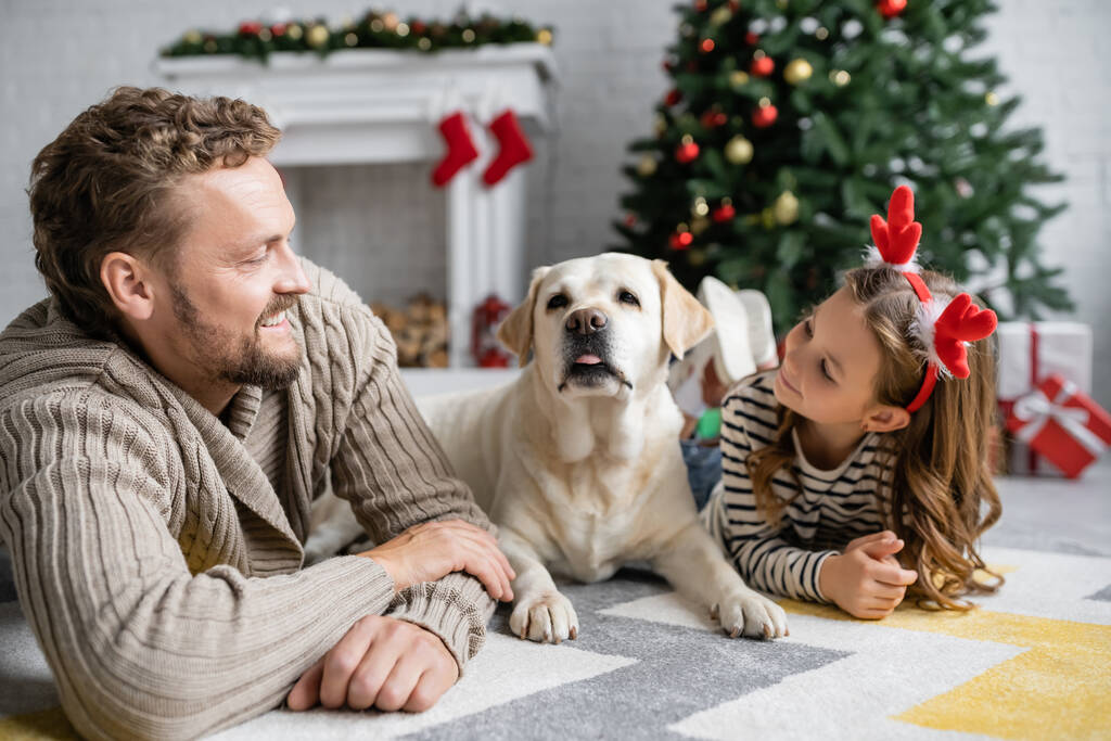 Hymyilevä isä katselee tytärtä joulun otsanauhalla lähellä labradoria kotona  - Valokuva, kuva