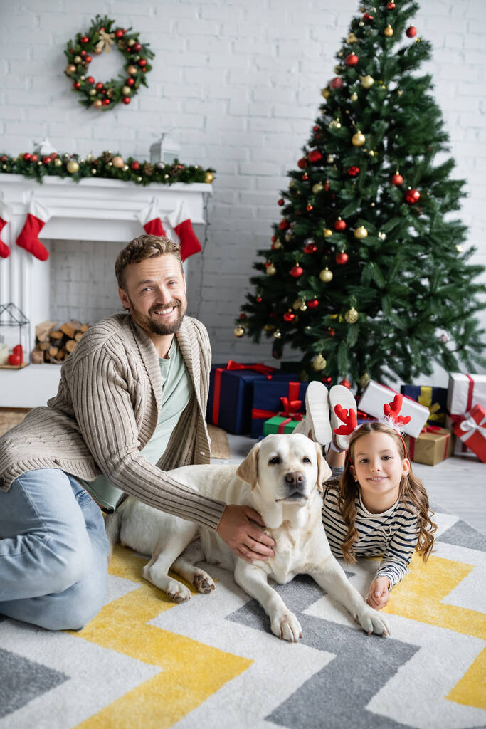 Father and child petting labrador near blurred christmas tree and gifts at home  - Photo, Image