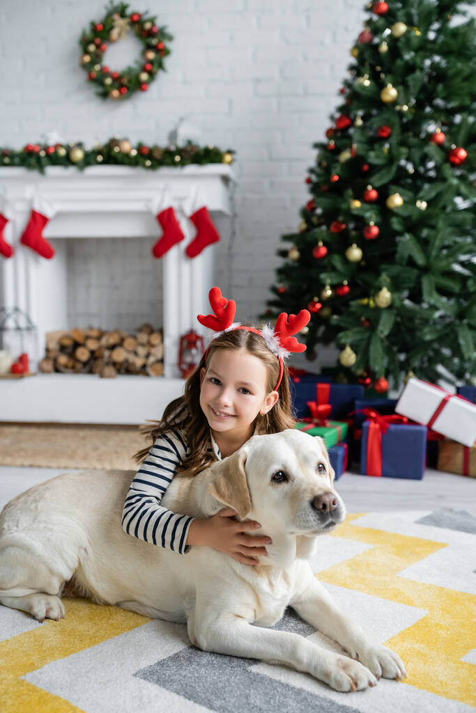 Vidám lány simogató labrador és a kamera közelében karácsonyfa a nappaliban  - Fotó, kép