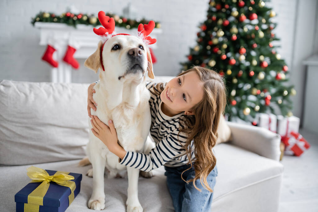 Хлопчик обіймає лабрадора з різдвяною пов'язкою біля присутніх на дивані вдома
  - Фото, зображення