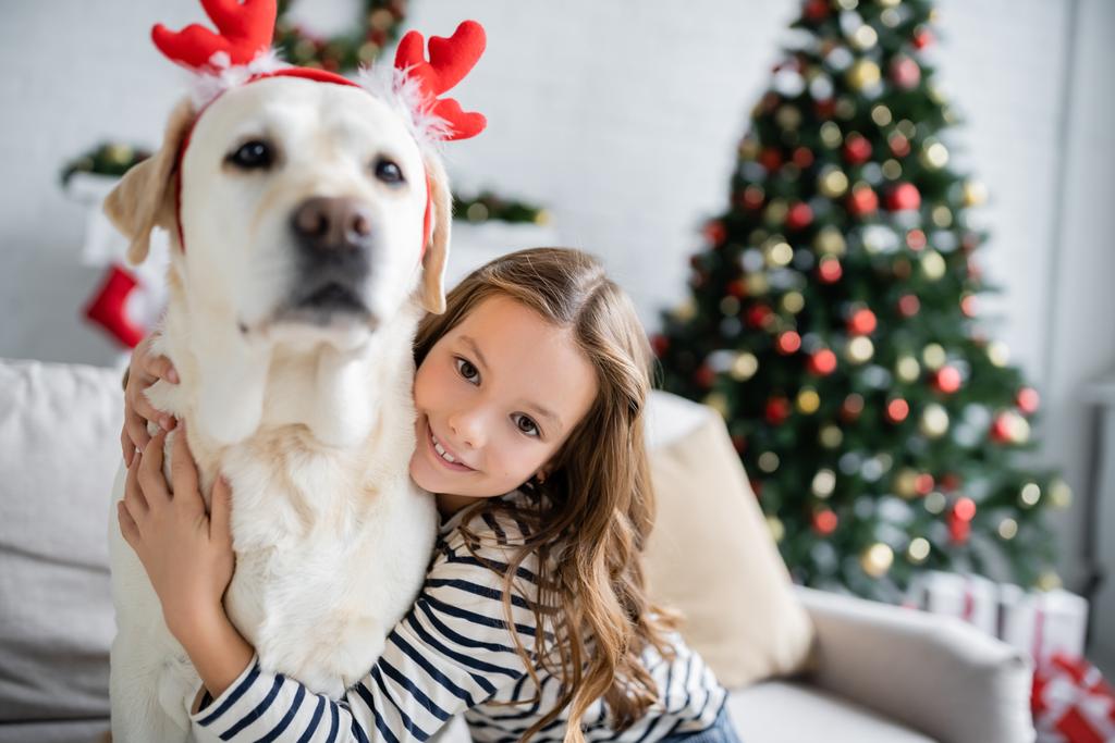 Chica sonriente abrazando labrador con diadema de Navidad y mirando a la cámara en la sala de estar  - Foto, Imagen