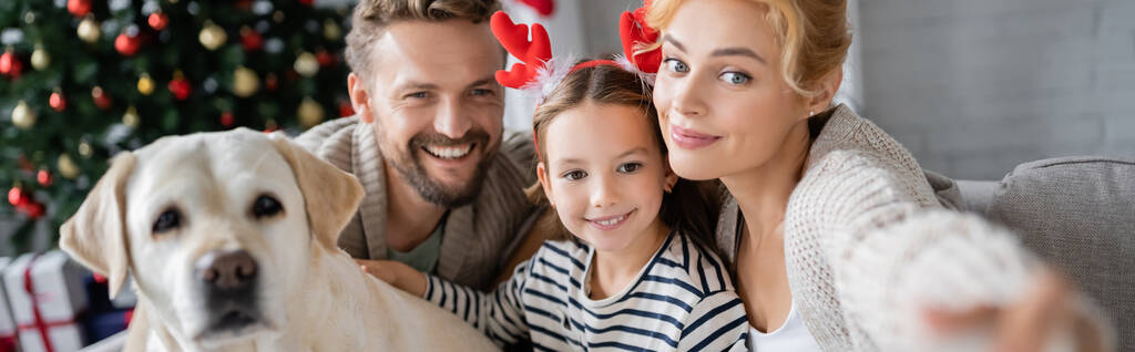 Χαμογελώντας οικογένεια και λαμπραντόρ κοιτάζοντας μακριά κατά τη διάρκεια των Χριστουγέννων στο σπίτι, πανό  - Φωτογραφία, εικόνα