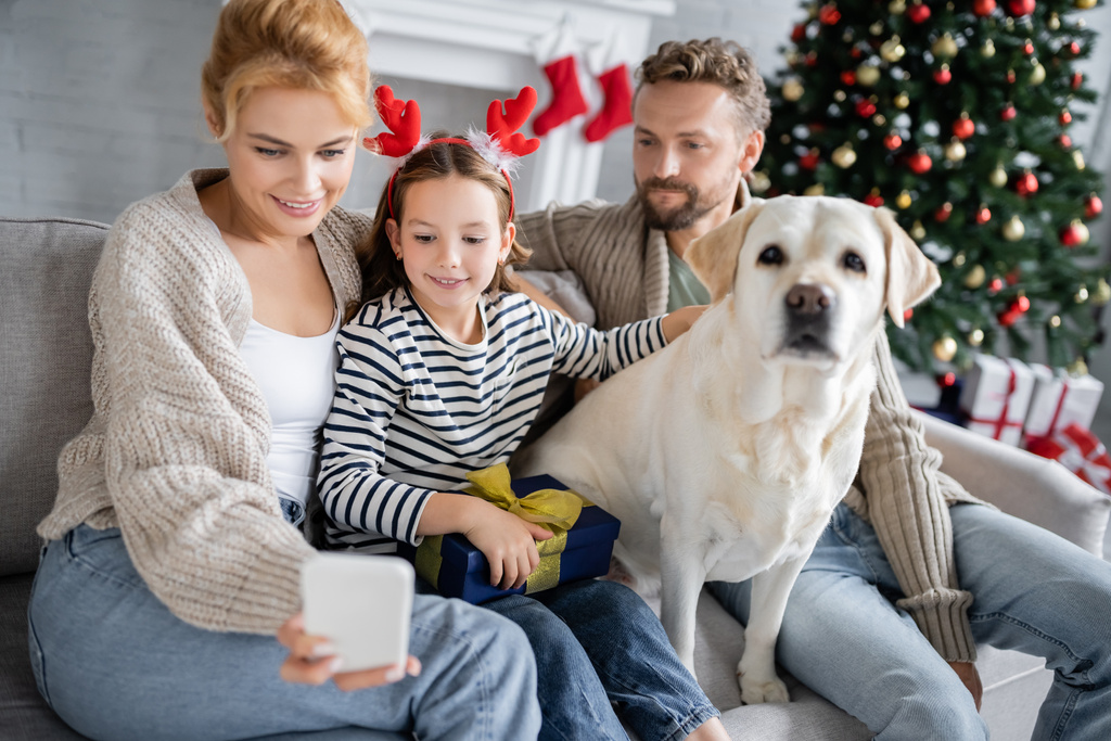 Mujer positiva usando celular cerca de hija sosteniendo regalo de Navidad y marido con labrador en casa  - Foto, imagen