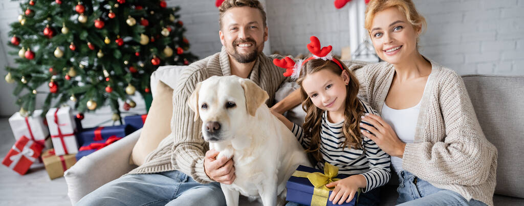 Fröhliche Familie mit Neujahrsgeschenk streichelt Labrador und schaut zu Hause in die Kamera, Banner  - Foto, Bild