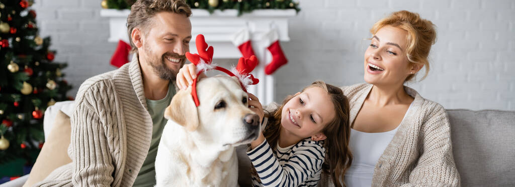 Famille positive portant bandeau de Noël sur labrador dans le salon, bannière  - Photo, image