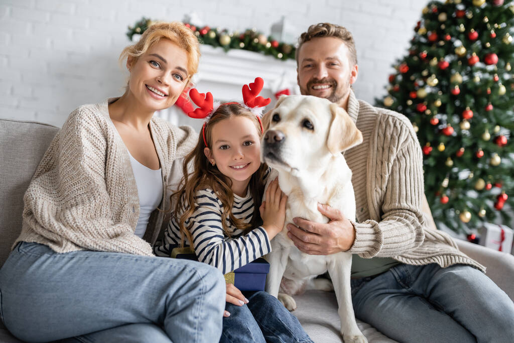 Sonrientes padres mirando a la cámara cerca de su hija con diadema de navidad abrazando a labrador en casa  - Foto, imagen
