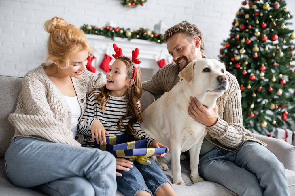 Girl opening christmas gift near parents and labrador at home  - Photo, Image