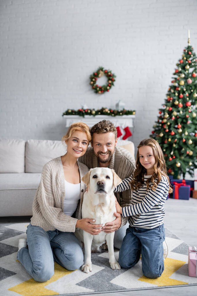 Pais felizes e menina olhando para a câmera perto labrador no tapete e árvore de natal borrada em casa  - Foto, Imagem