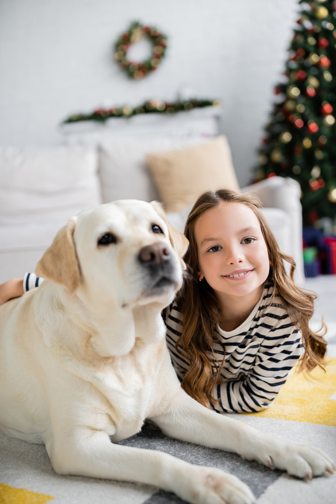 Lächelndes Mädchen umarmt Labrador, während es zu Neujahr auf dem Boden liegt  - Foto, Bild