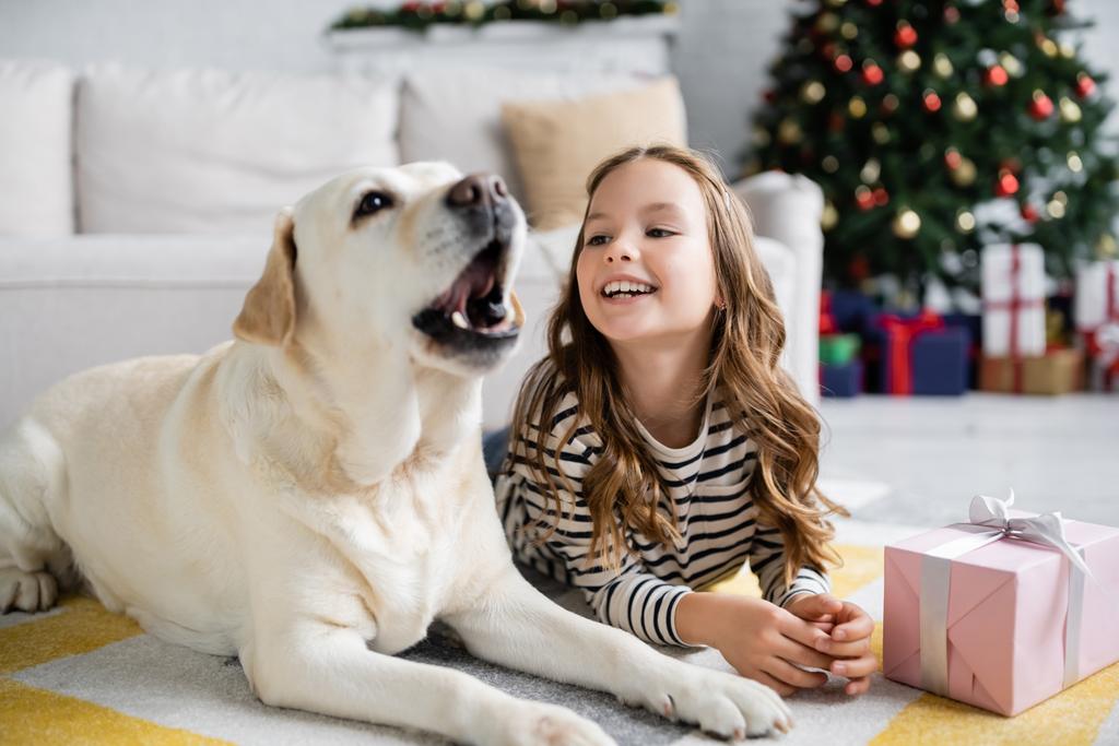 Fröhliches Mädchen sieht Labrador-Hund kurz vor Neujahr zu Hause auf dem Boden  - Foto, Bild