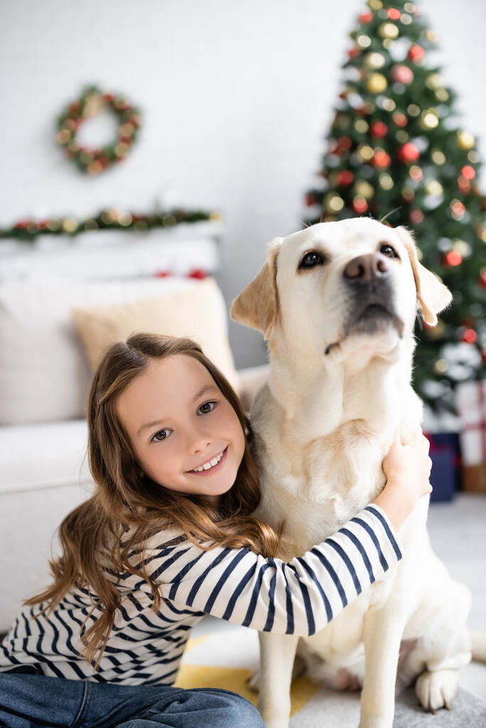 Dziewczyna uśmiecha się do kamery, przytulając labradora w pobliżu rozmytej choinki w domu  - Zdjęcie, obraz