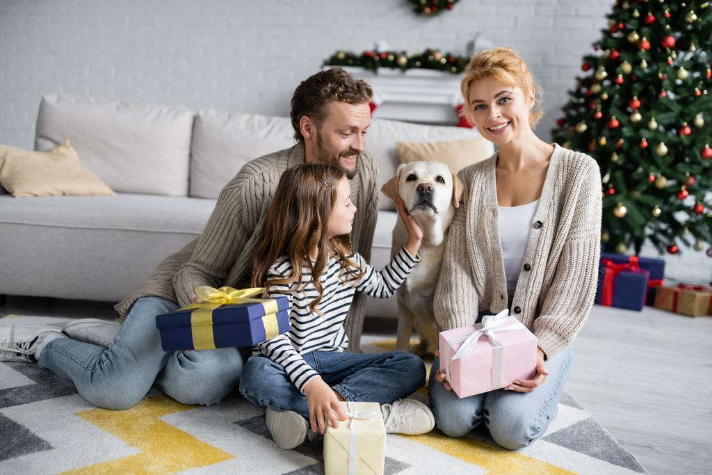 Femme souriante tenant cadeau de Noël tandis que la famille caresse labrador à la maison  - Photo, image